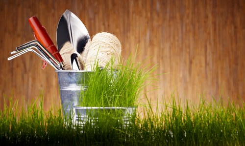 The image depicts a close-up of a garden scene with a metal container holding various gardening tools, including a trowel, a rake, and a pair of pruning shears, along with a ball of twine. In the foreground, there is vibrant green grass, suggesting a well-maintained lawn. The background features a warm wooden surface, possibly a garden fence or wall, providing a natural backdrop that complements the outdoor setting. This scene reflects outdoor gardening activities typical in residential gardens within Marylebone, showcasing tools and plant elements that might be used in regular lawn and hedge maintenance, consistent with services offered by Hedge Trimming Marylebone. The lighting appears natural, indicating a bright, clear day, ideal for garden work. The composition emphasizes tools ready for use, highlighting the importance of proper garden care and maintenance in an outdoor space, aligning with professional gardening and landscaping services in the London area, including Marylebone postcode zones.