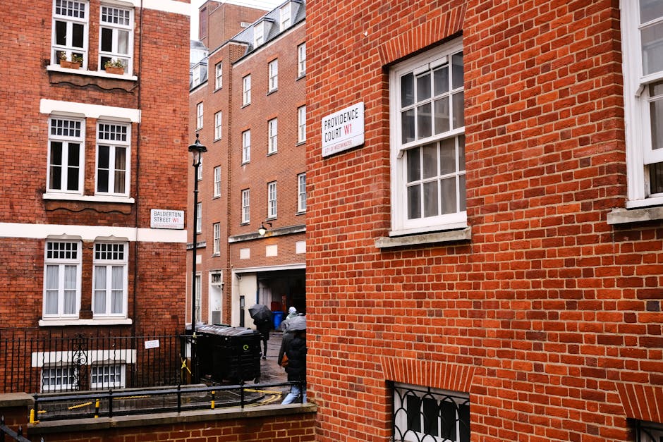 A narrow urban street corner featuring red brick residential buildings with white-framed sash windows. The foreground shows a small garden area enclosed by a black iron fence, with parts of paved path