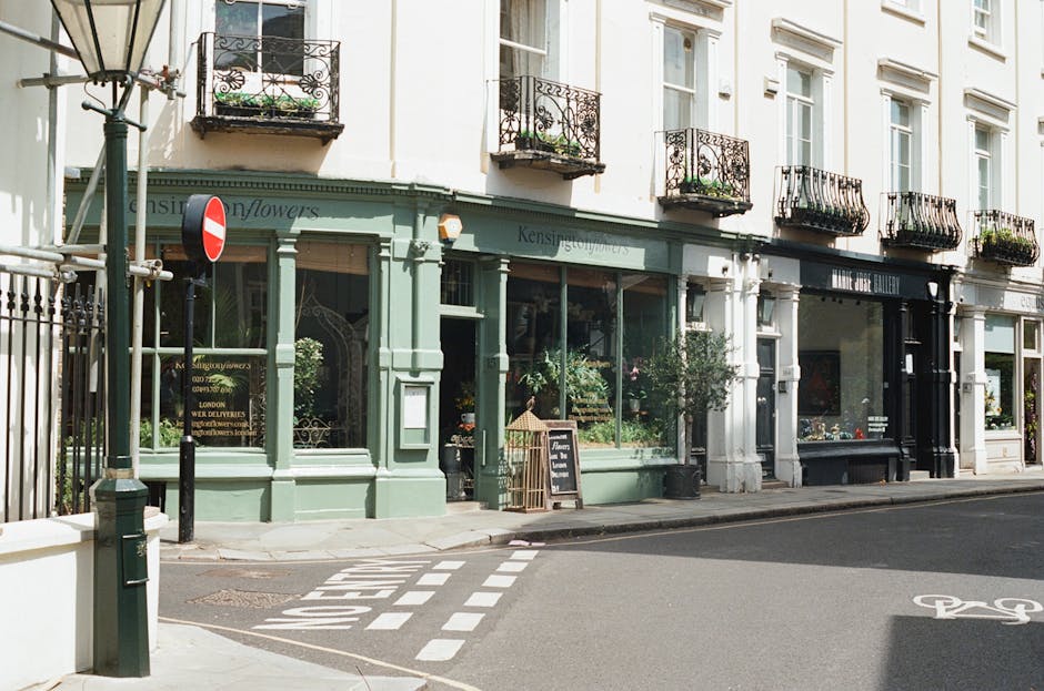 A narrow urban street corner featuring red brick residential buildings with white-framed sash windows. The foreground shows a small garden area enclosed by a black iron fence, with parts of paved path