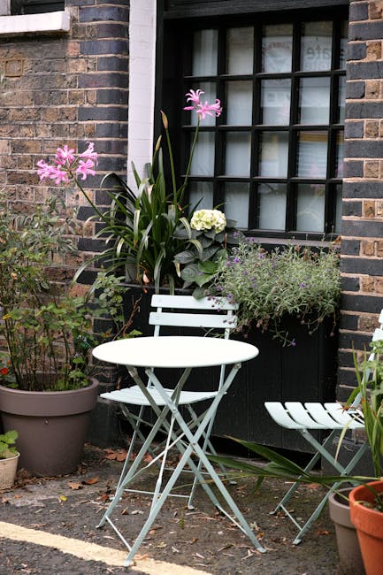 A cozy small garden situated against the brick exterior of a building, featuring a variety of lush green plants and foliage. In the foreground, there are potted plants with fern-like leaves and a smal